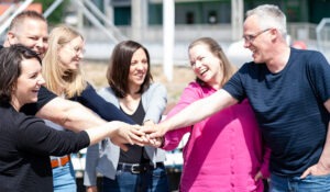Eine Gruppe von sechs fröhlichen Menschen steht im Kreis und legt ihre Hände übereinander als Zeichen des Teamgeists.