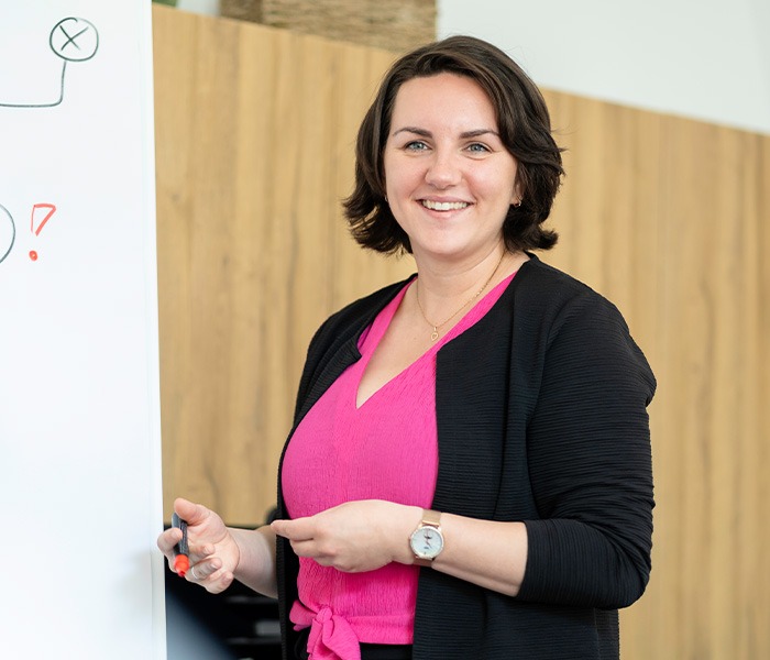 Eine Frau mit kurzen, dunklen Haaren steht lächelnd neben einem Whiteboard und hält einen Stift in der Hand; sie trägt eine schwarze Jacke über einem pinken Oberteil.