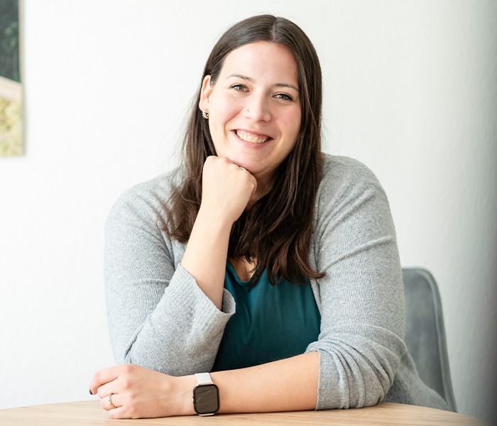Eine Frau mit langen dunklen Haaren und einem freundlichen Lächeln stützt ihren Kopf auf ihre Hand und trägt eine graue Strickjacke sowie eine Armbanduhr, während sie an einem Tisch sitzt.