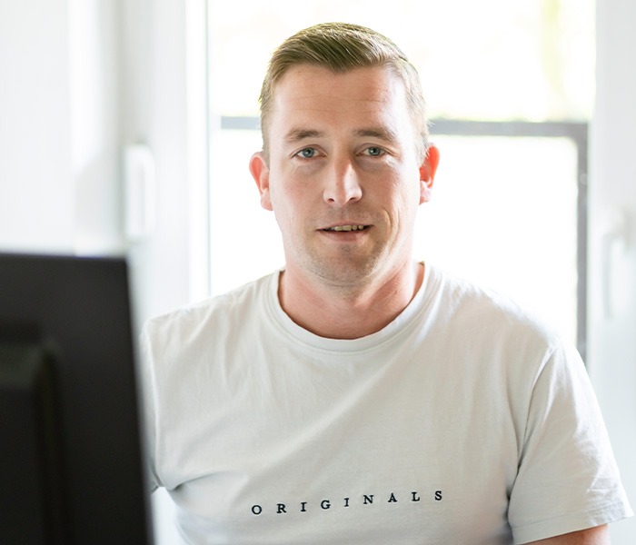 Ein Mann in einem weißen T-Shirt mit der Aufschrift "Originals" sitzt vor einem Computerbildschirm und schaut in die Kamera.