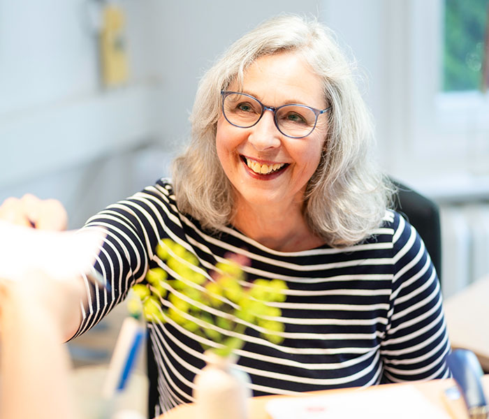 Eine lächelnde Frau mit Brille und grauem Haar sitzt an einem Tisch und trägt ein gestreiftes Oberteil, während sie einer anderen Person etwas überreicht.